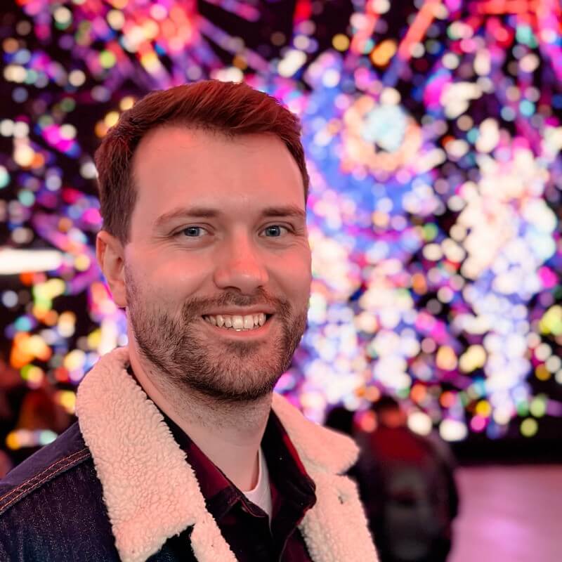 David Cox's profile photo, cropped below the shoulders. David beams a smile while standing in front of a myriad of blurred purple, yellow, blue and magenta lights.